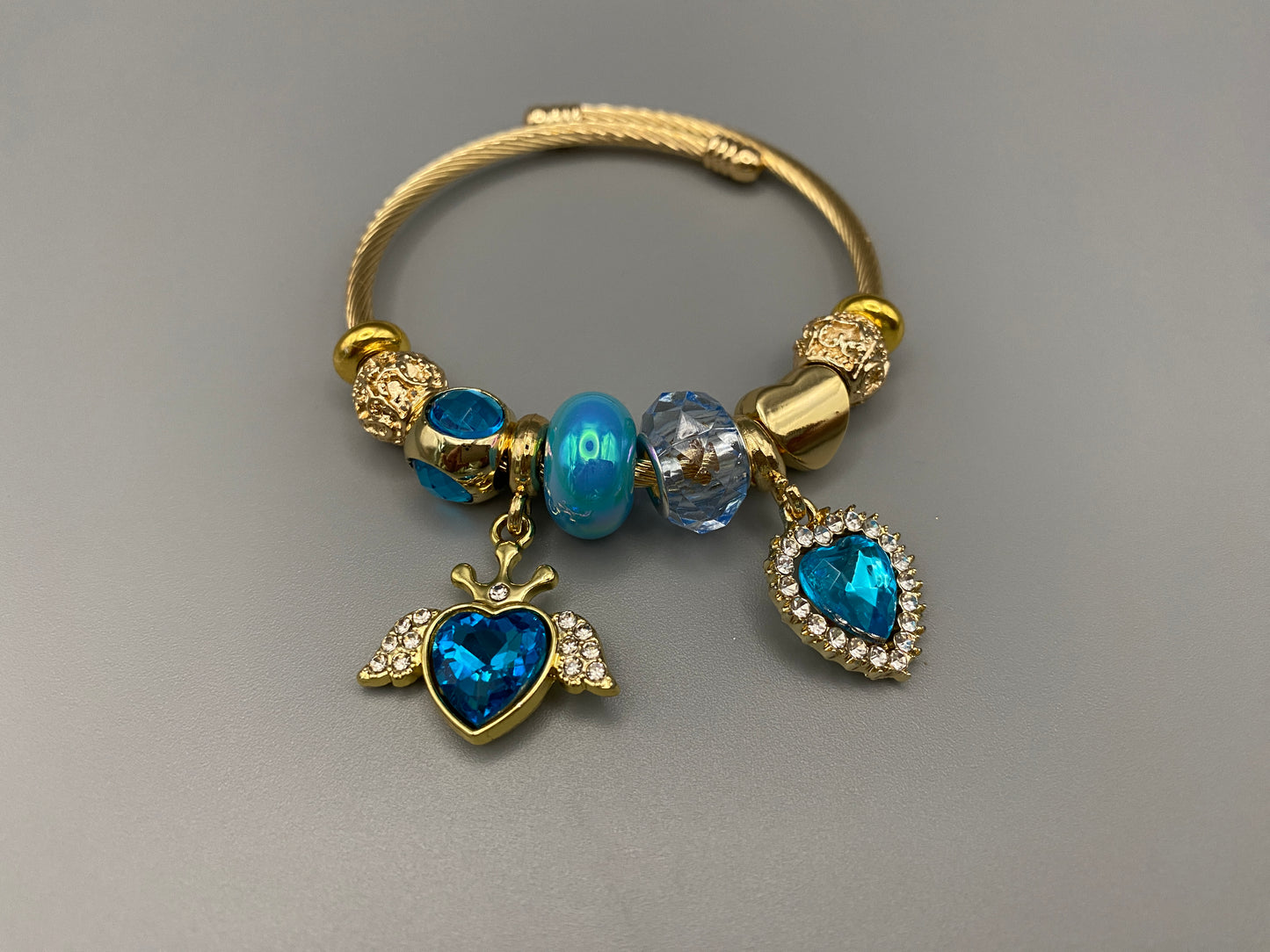 “Belle Blue Charm Bracelet with gold-plated finish, featuring heart-cut crystals and rhinestones. Made from durable stainless steel with playful charms and micro insert inlay technology.”