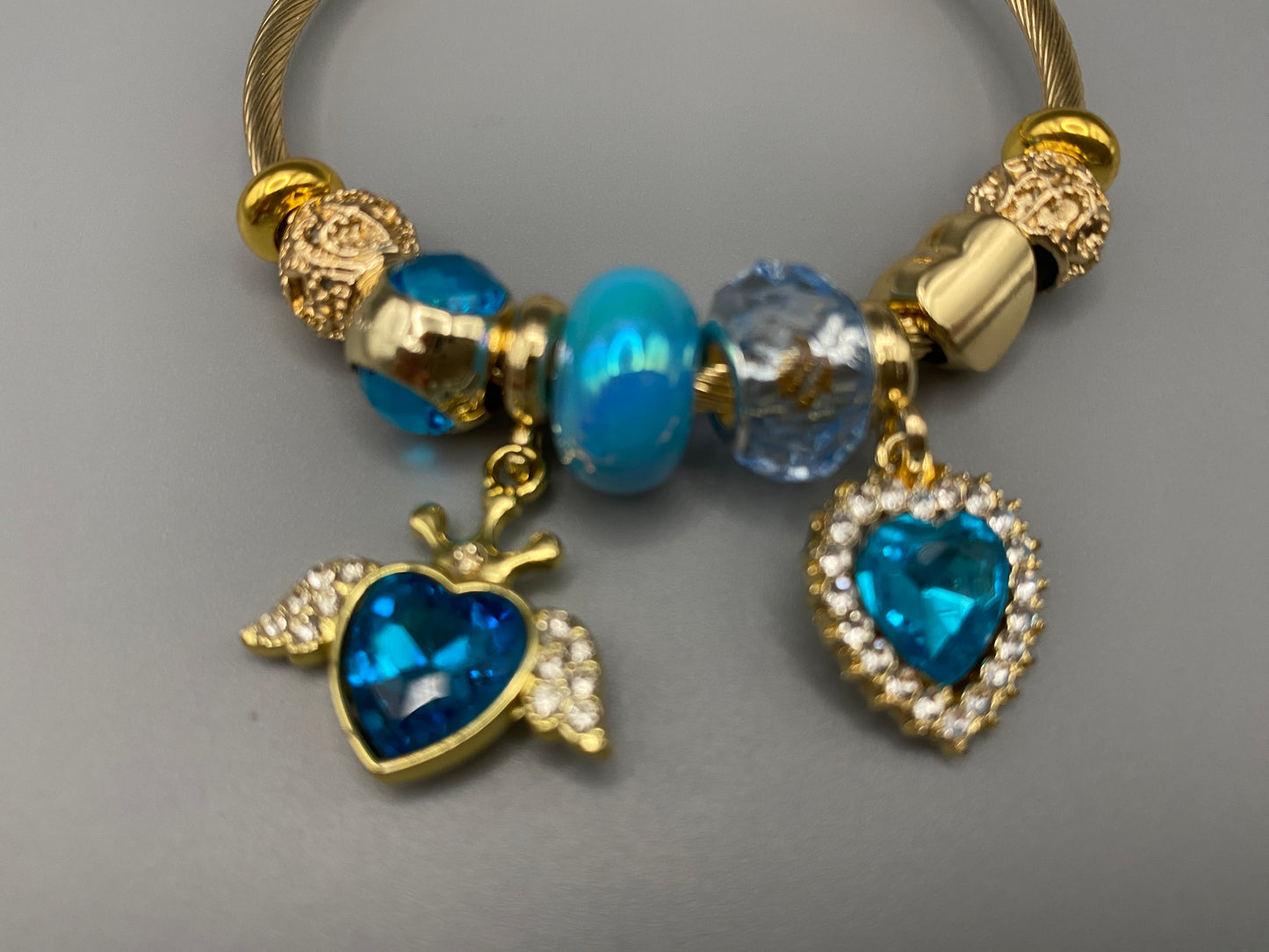 “Belle Blue Charm Bracelet with gold-plated finish, featuring heart-cut crystals and rhinestones. Made from durable stainless steel with playful charms and micro insert inlay technology.”