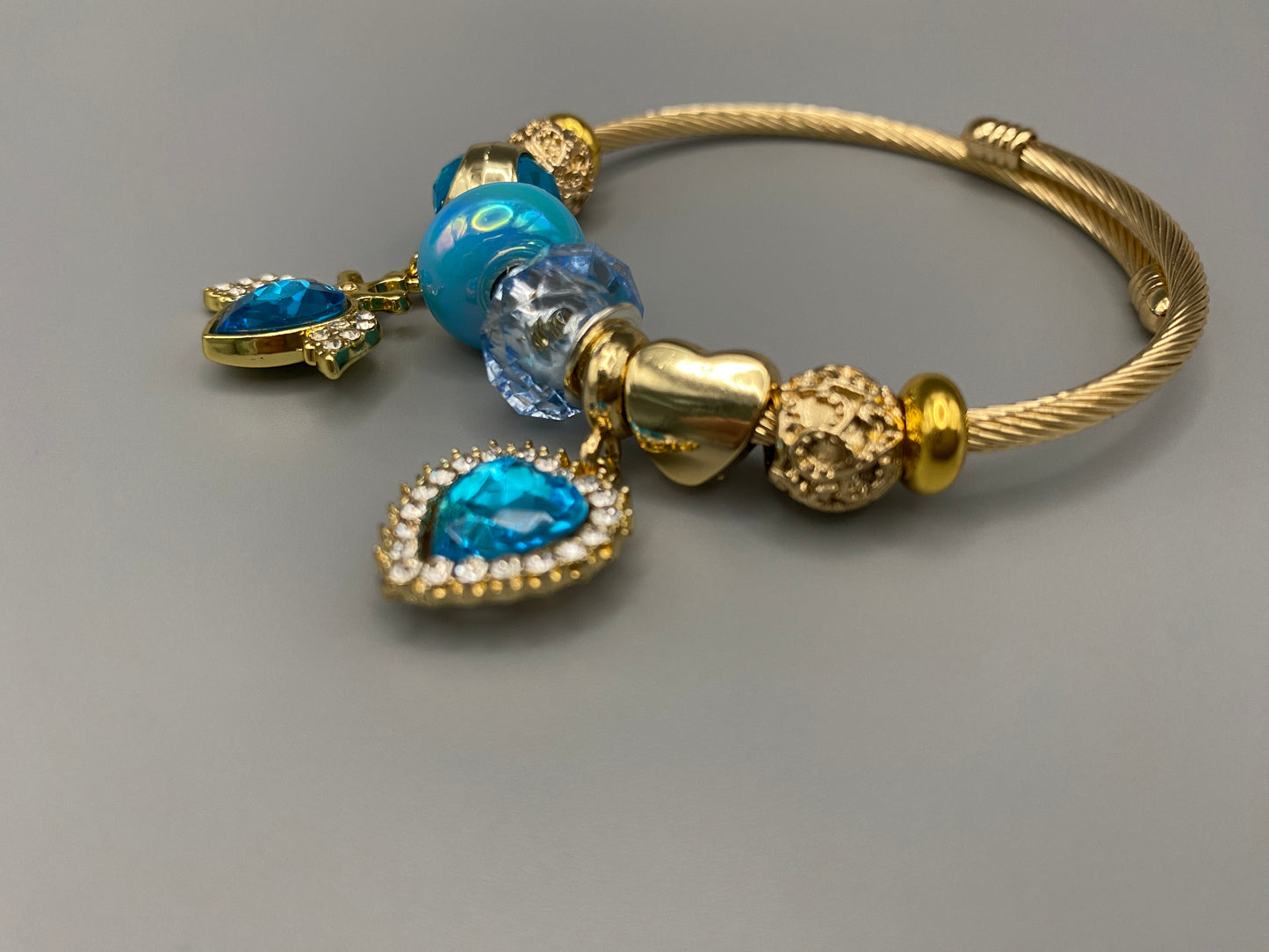 “Belle Blue Charm Bracelet with gold-plated finish, featuring heart-cut crystals and rhinestones. Made from durable stainless steel with playful charms and micro insert inlay technology.”