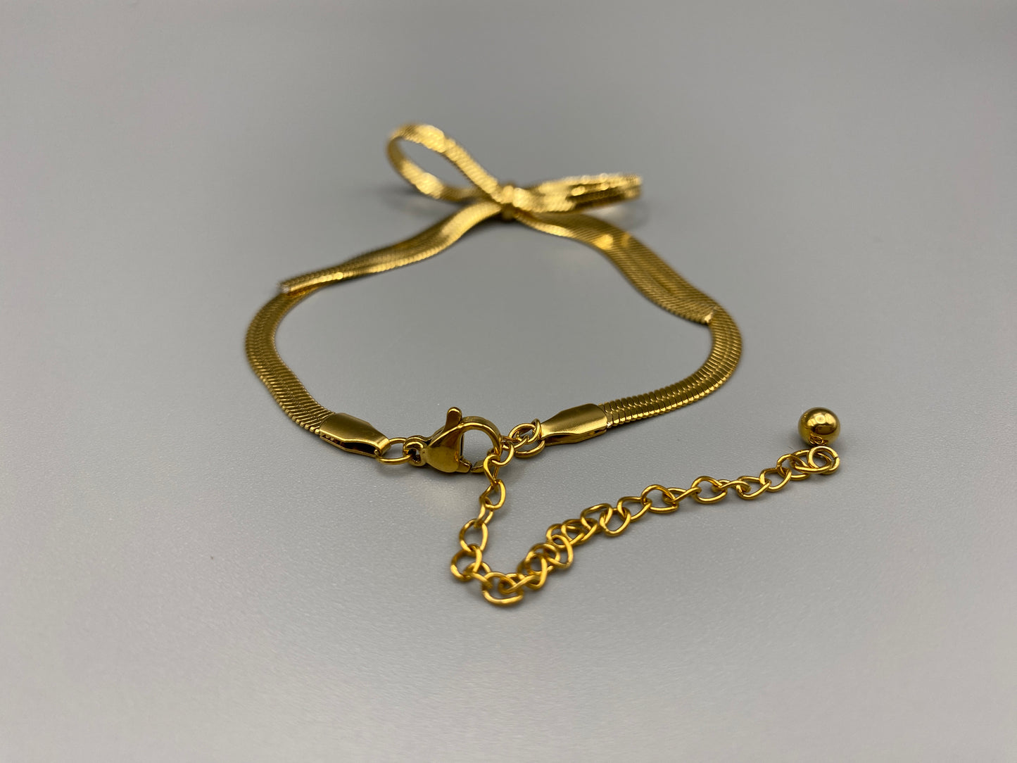 “The Celest Angel Bow Bracelet in gold features a delicate bow design. It is made of stainless steel for durability and tarnish resistance. The bracelet measures 18 cm with a 3 cm extender for an adjustable fit.”