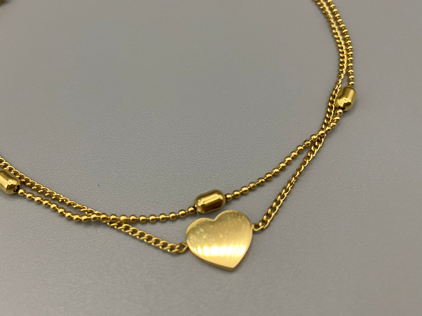 “Cora Twin Chain Heart and Bead Bracelet in gold, featuring a romantic twin chain design with heart and bead accents. 18K gold plated, made from durable titanium steel with PVD plating. Measures 17 cm with a 3 cm extender for adjustable fit.”