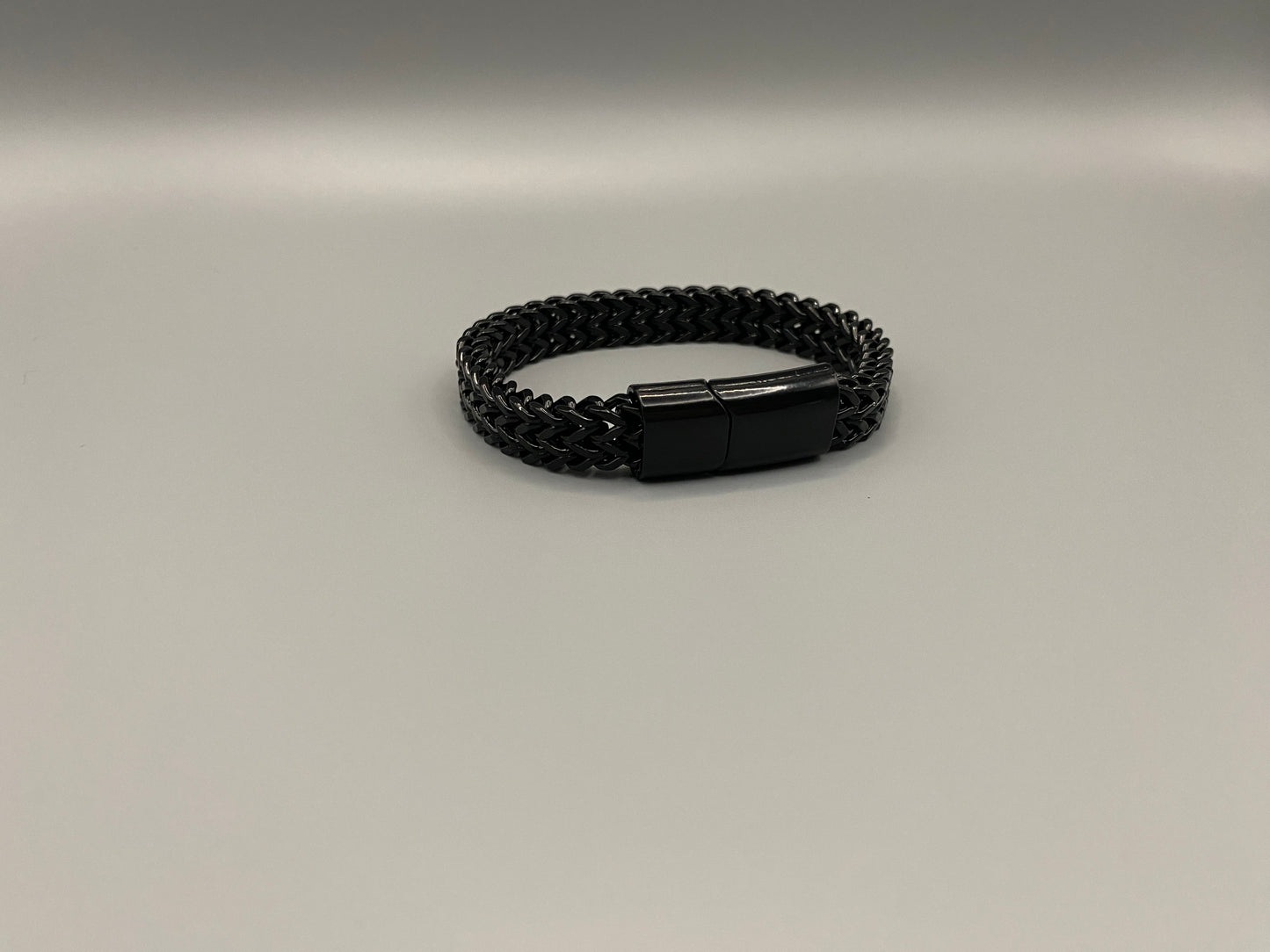 “A men’s bracelet with a chain and link design, made from shiny titanium steel. The bracelet features a geometric pattern and a smart magnet button clasp. It is available in silver, gold, and black colors, and measures 19-23 cm in length.”