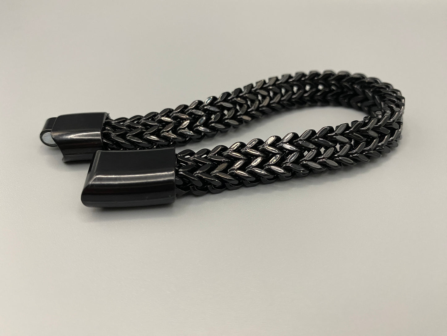 “A men’s bracelet with a chain and link design, made from shiny titanium steel. The bracelet features a geometric pattern and a smart magnet button clasp. It is available in silver, gold, and black colors, and measures 19-23 cm in length.”