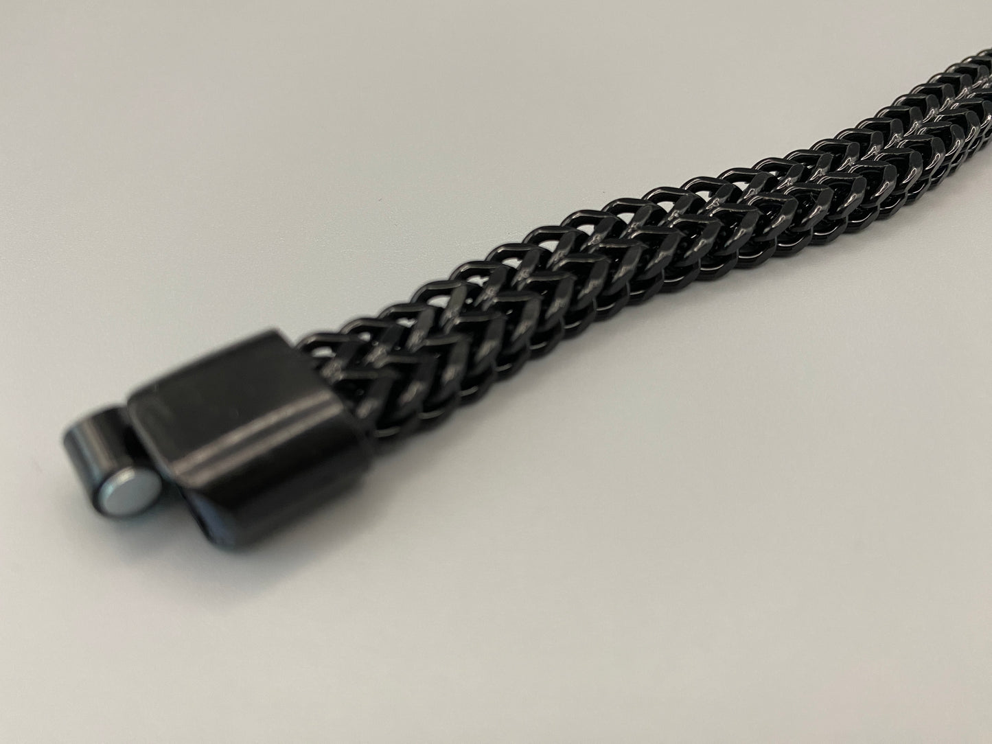 “A men’s bracelet with a chain and link design, made from shiny titanium steel. The bracelet features a geometric pattern and a smart magnet button clasp. It is available in silver, gold, and black colors, and measures 19-23 cm in length.”