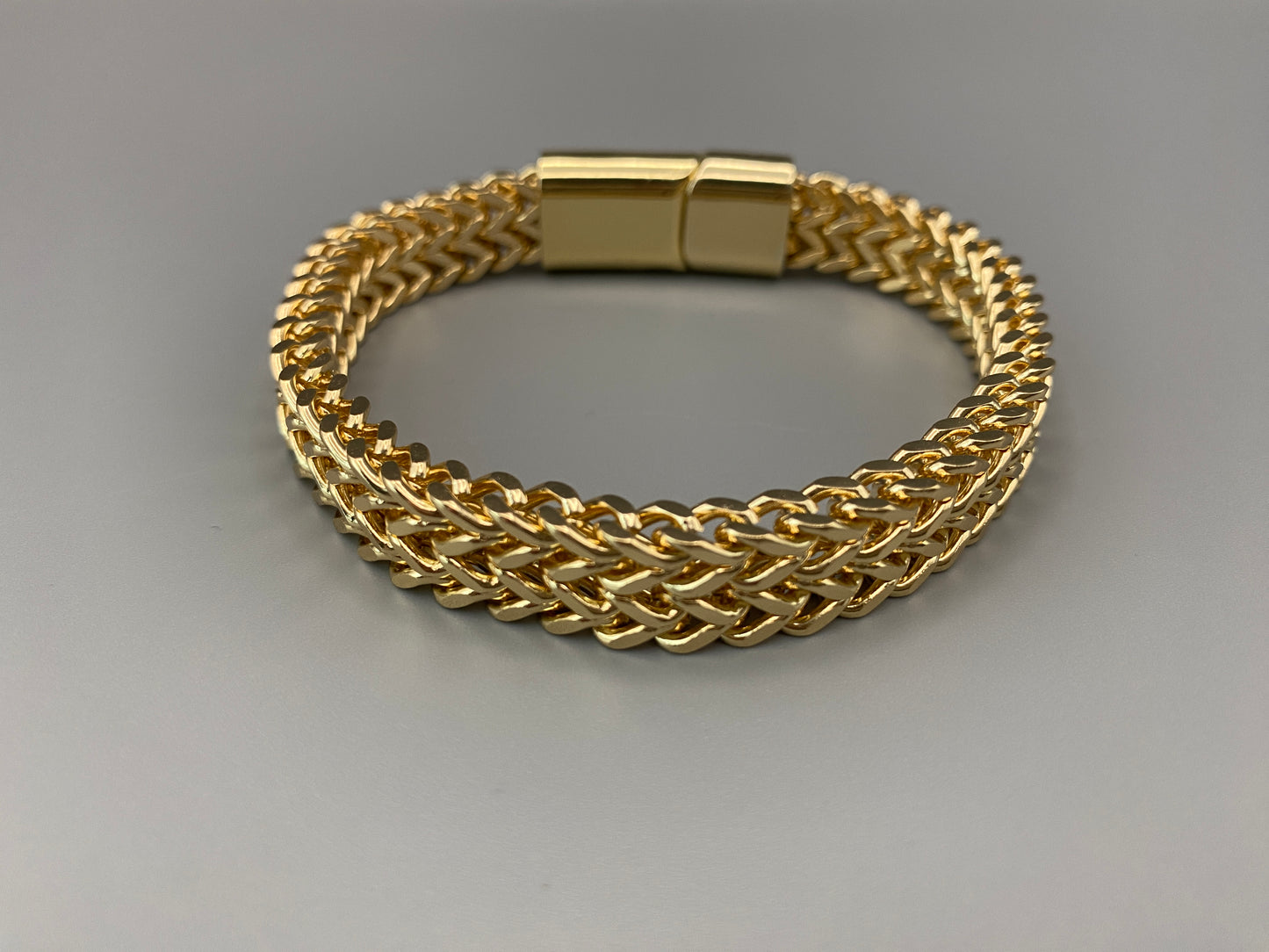 “A men’s bracelet with a chain and link design, made from shiny titanium steel. The bracelet features a geometric pattern and a smart magnet button clasp. It is available in silver, gold, and black colors, and measures 19-23 cm in length.”