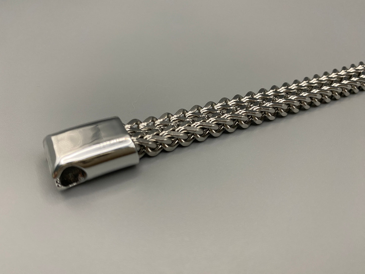 “A men’s bracelet with a chain and link design, made from shiny titanium steel. The bracelet features a geometric pattern and a smart magnet button clasp. It is available in silver, gold, and black colors, and measures 19-23 cm in length.”