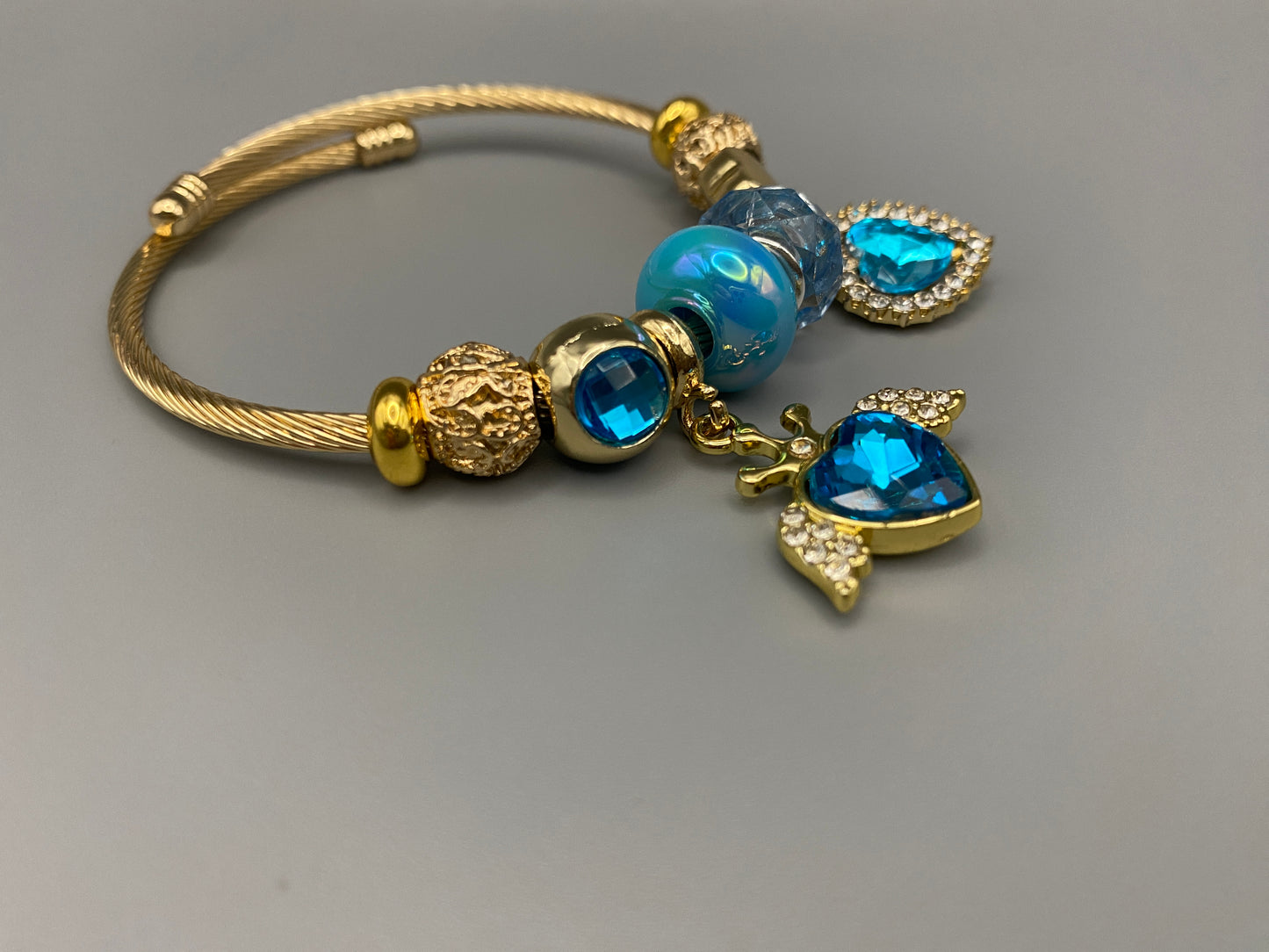 “Belle Blue Charm Bracelet with gold-plated finish, featuring heart-cut crystals and rhinestones. Made from durable stainless steel with playful charms and micro insert inlay technology.”