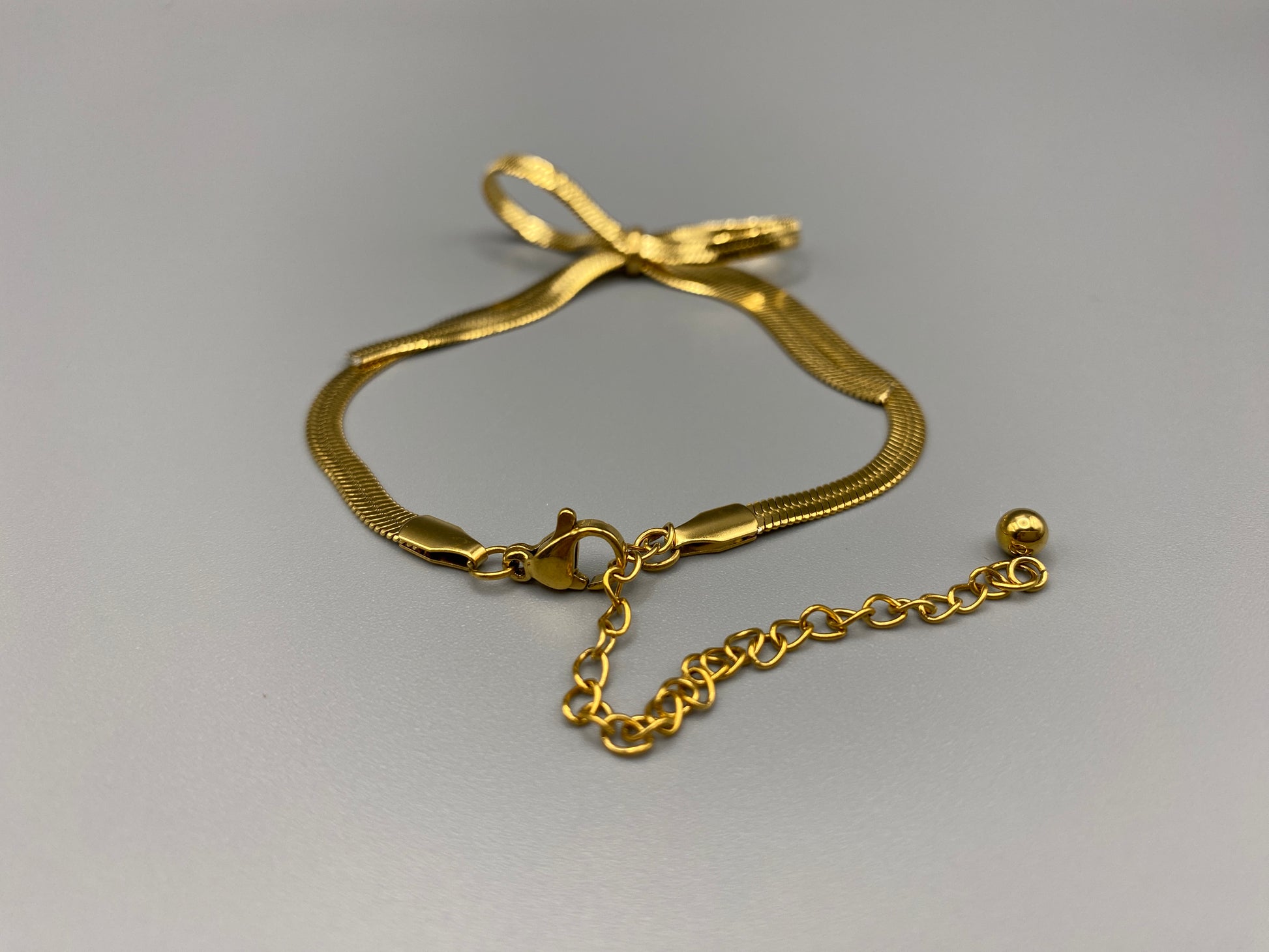 “The Celest Angel Bow Bracelet in gold features a delicate bow design. It is made of stainless steel for durability and tarnish resistance. The bracelet measures 18 cm with a 3 cm extender for an adjustable fit.”