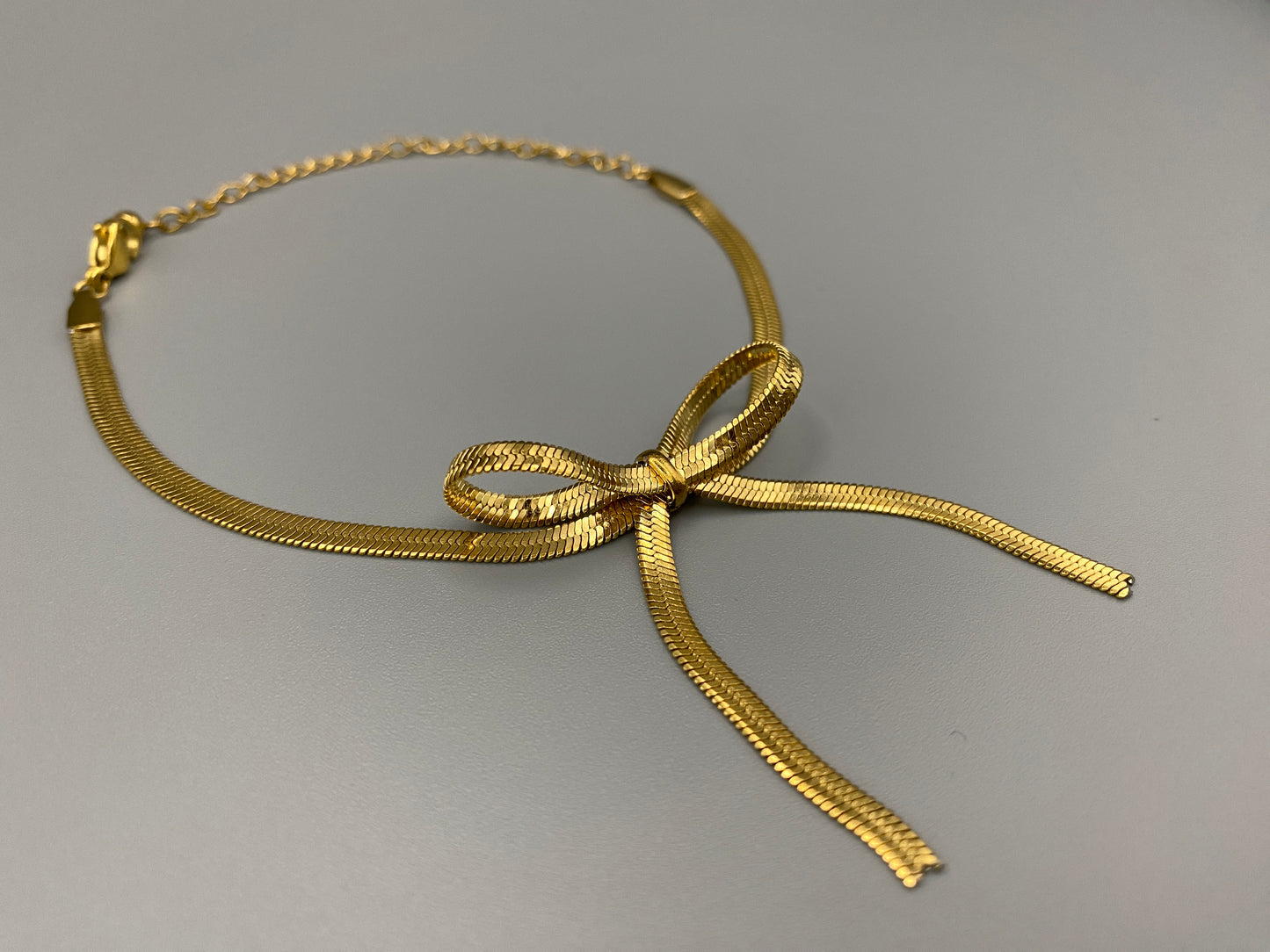 “The Celest Angel Bow Bracelet in gold features a delicate bow design. It is made of stainless steel for durability and tarnish resistance. The bracelet measures 18 cm with a 3 cm extender for an adjustable fit.”