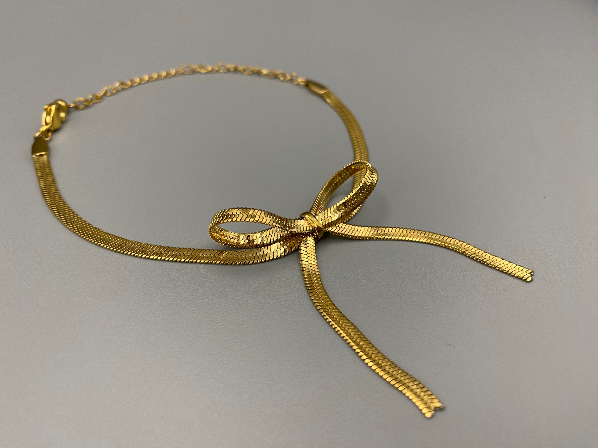 “The Celest Angel Bow Bracelet in gold features a delicate bow design. It is made of stainless steel for durability and tarnish resistance. The bracelet measures 18 cm with a 3 cm extender for an adjustable fit.”