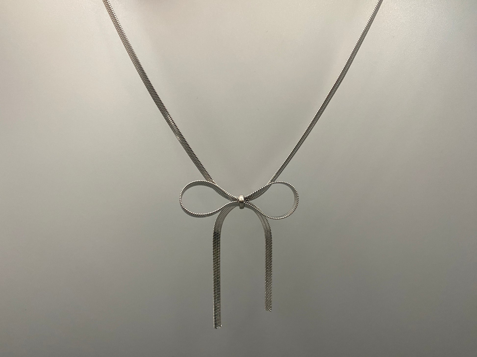“Celest Angel Bow Pendant Chain in silver with a sophisticated bow pendant design. Made from durable stainless steel, tarnish-free. Measures 35 cm with a 5 cm extender for adjustable fit.”