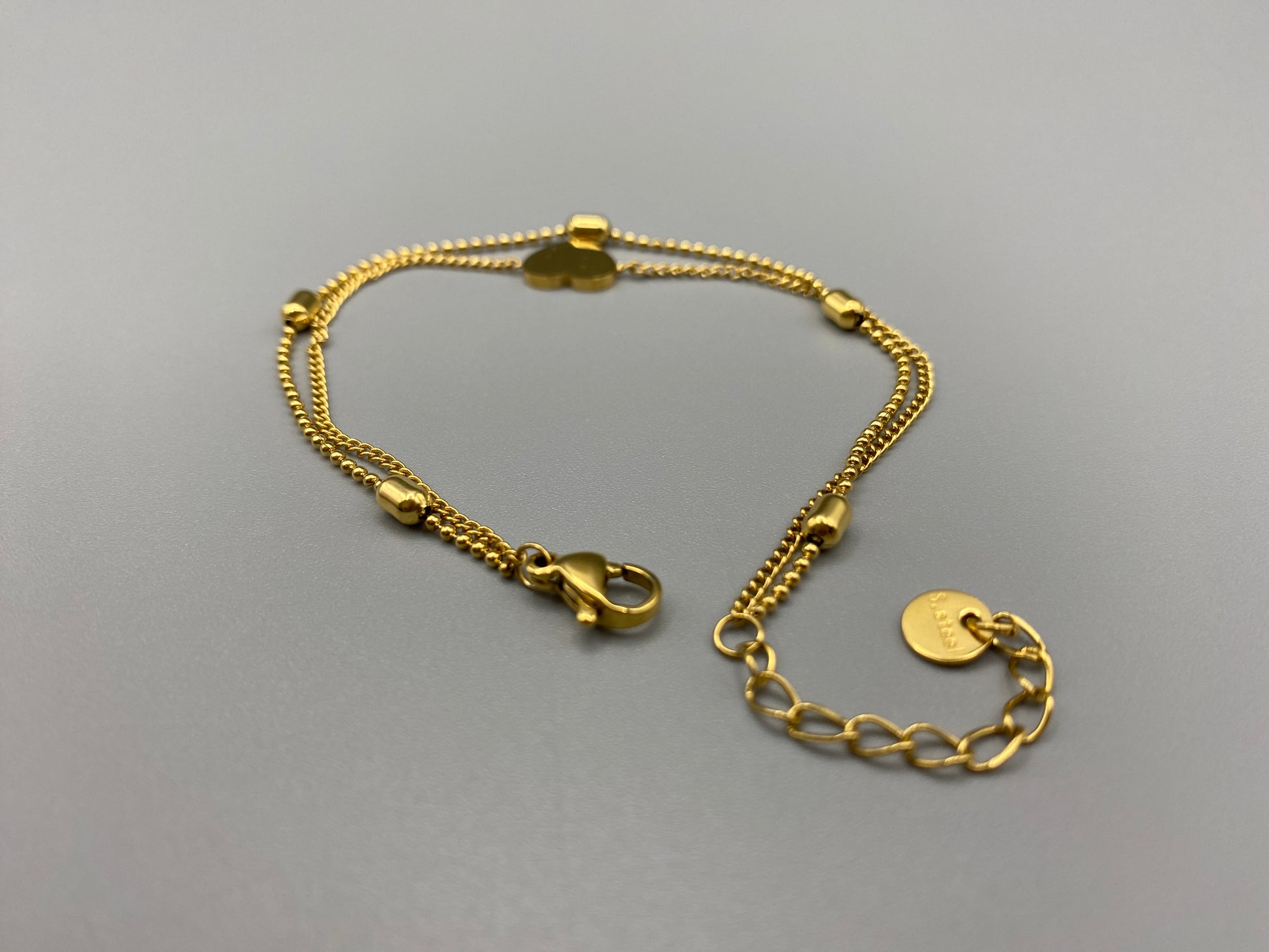 “Cora Twin Chain Heart and Bead Bracelet in gold, featuring a romantic twin chain design with heart and bead accents. 18K gold plated, made from durable titanium steel with PVD plating. Measures 17 cm with a 3 cm extender for adjustable fit.”