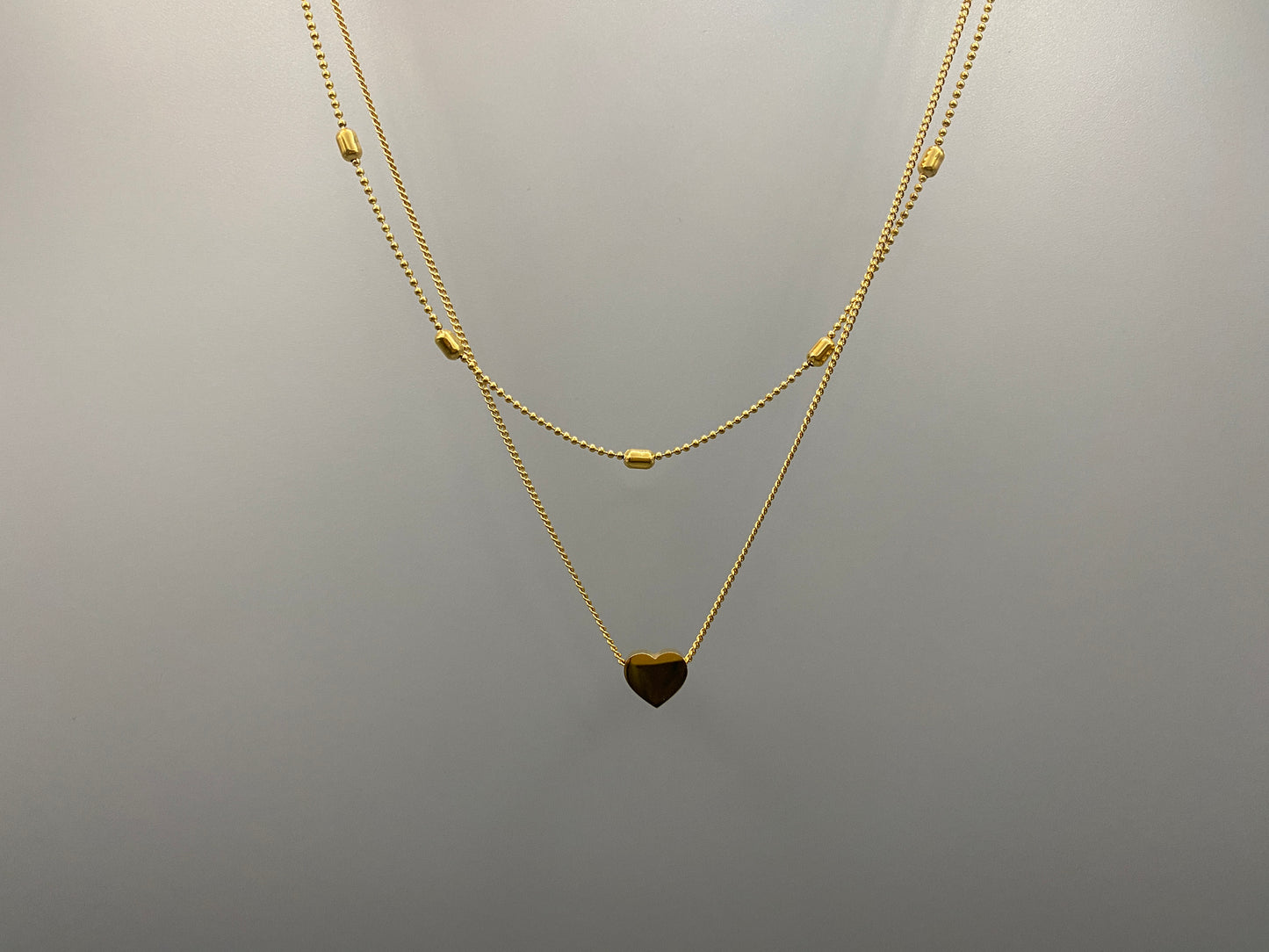 Cora, This exquisite necklace is crafted from luxurious 18K gold, featuring a unique twin chain design. One chain is adorned with delicate beads, while the other showcases a charming heart-shaped pendant.