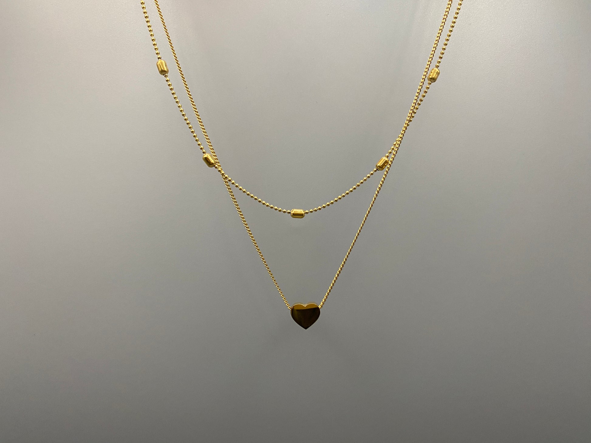 Cora, This exquisite necklace is crafted from luxurious 18K gold, featuring a unique twin chain design. One chain is adorned with delicate beads, while the other showcases a charming heart-shaped pendant.