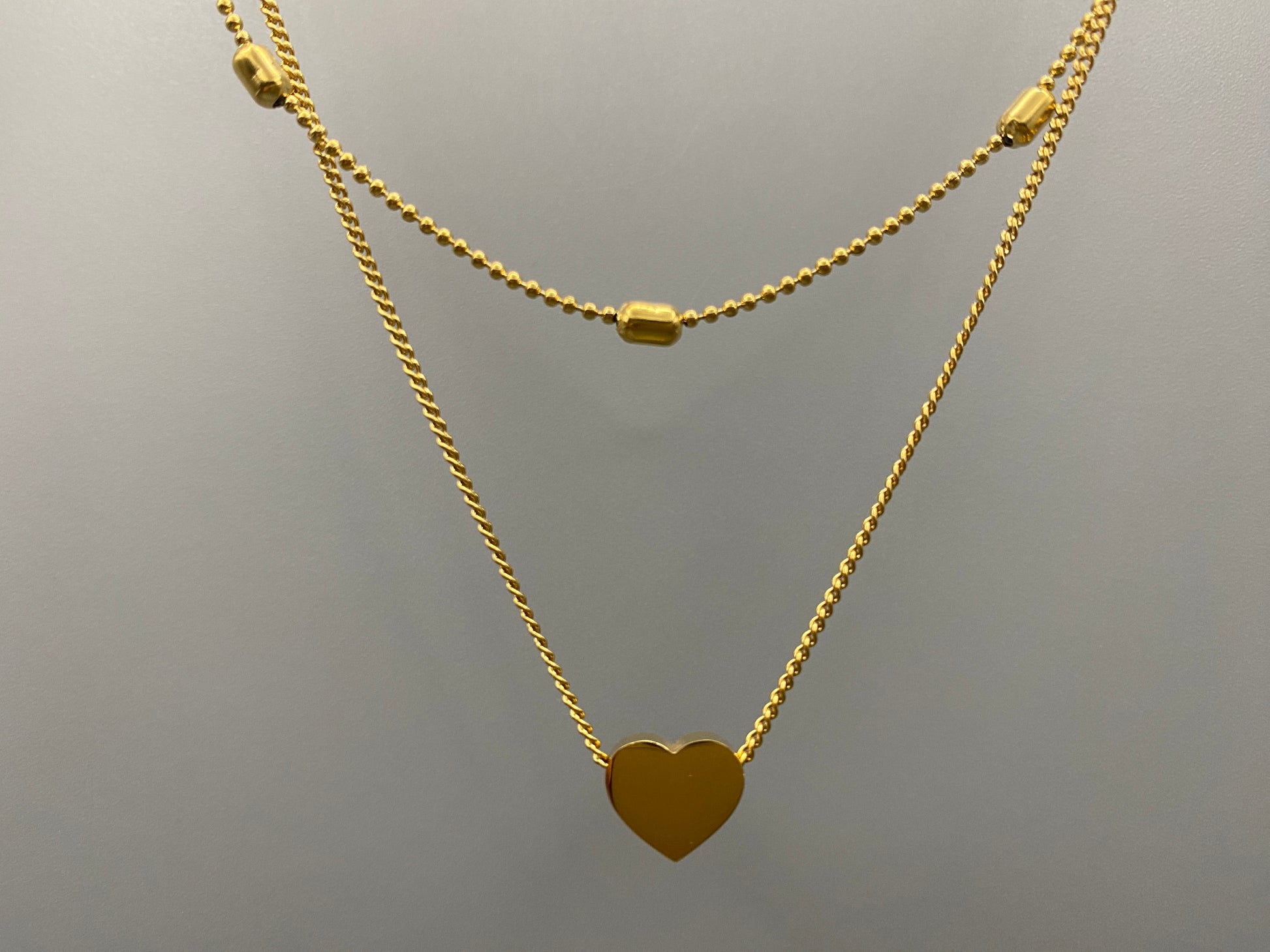 Cora, This exquisite necklace is crafted from luxurious 18K gold, featuring a unique twin chain design. One chain is adorned with delicate beads, while the other showcases a charming heart-shaped pendant.