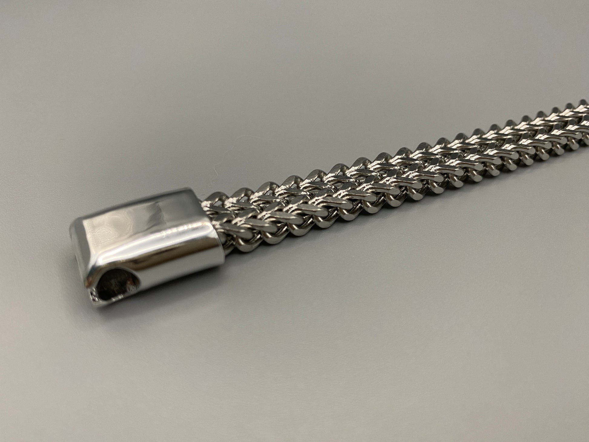 “A men’s bracelet with a chain and link design, made from shiny titanium steel. The bracelet features a geometric pattern and a smart magnet button clasp. It is available in silver, gold, and black colors, and measures 19-23 cm in length.”