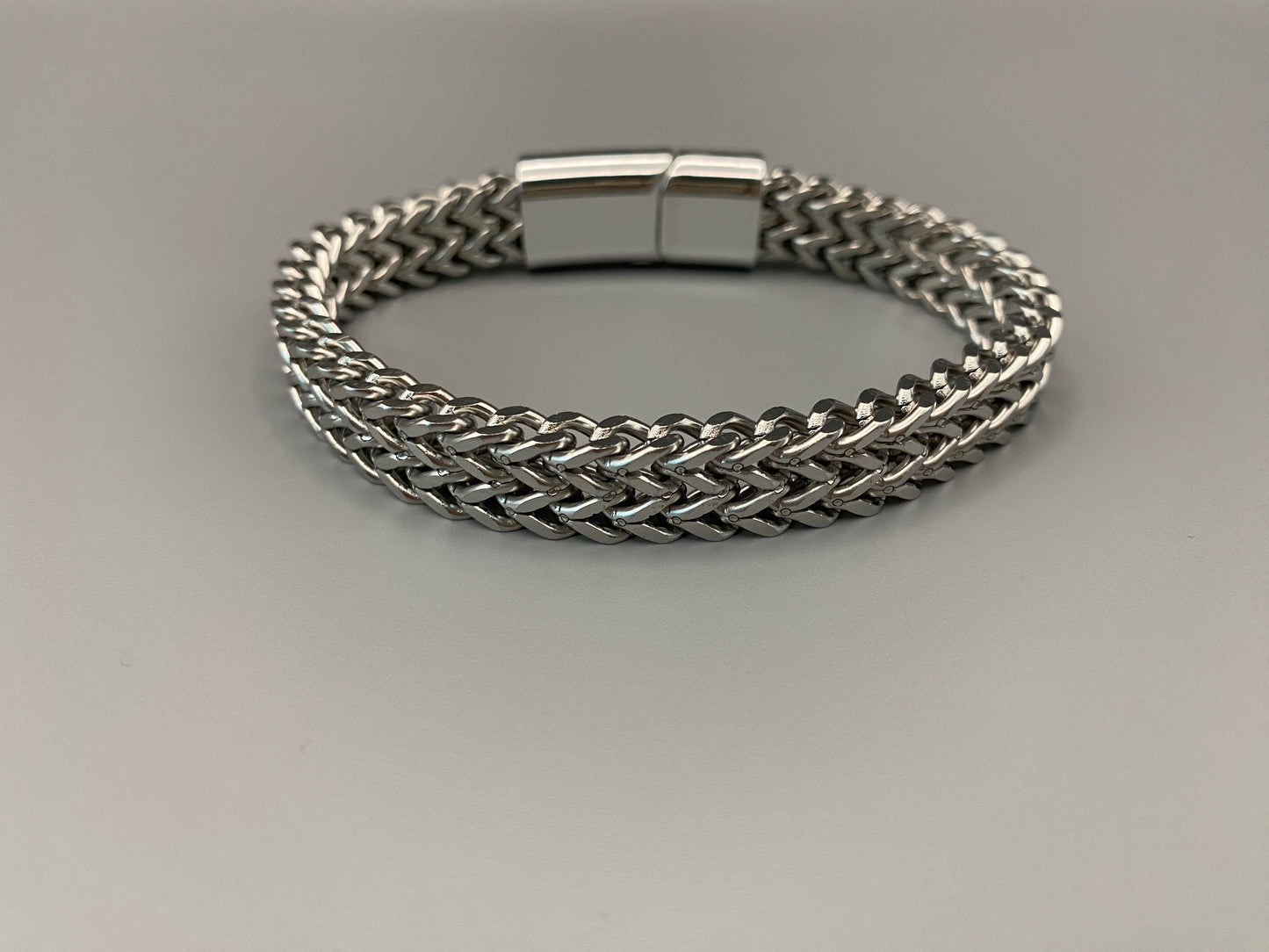 “A men’s bracelet with a chain and link design, made from shiny titanium steel. The bracelet features a geometric pattern and a smart magnet button clasp. It is available in silver, gold, and black colors, and measures 19-23 cm in length.”