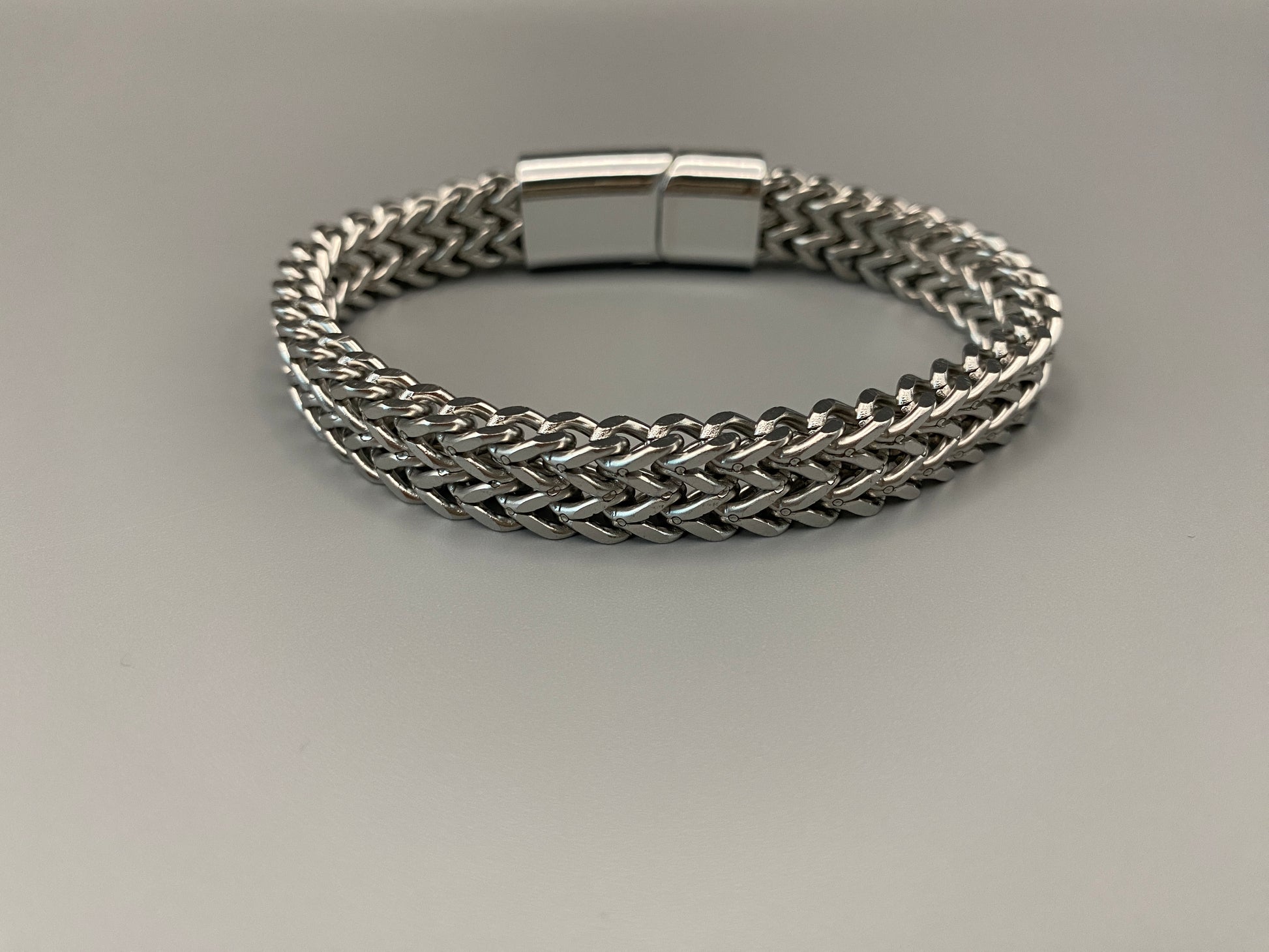 “A men’s bracelet with a chain and link design, made from shiny titanium steel. The bracelet features a geometric pattern and a smart magnet button clasp. It is available in silver, gold, and black colors, and measures 19-23 cm in length.”
