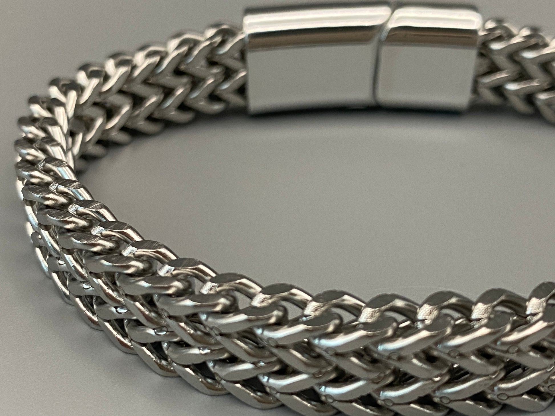 “A men’s bracelet with a chain and link design, made from shiny titanium steel. The bracelet features a geometric pattern and a smart magnet button clasp. It is available in silver, gold, and black colors, and measures 19-23 cm in length.”