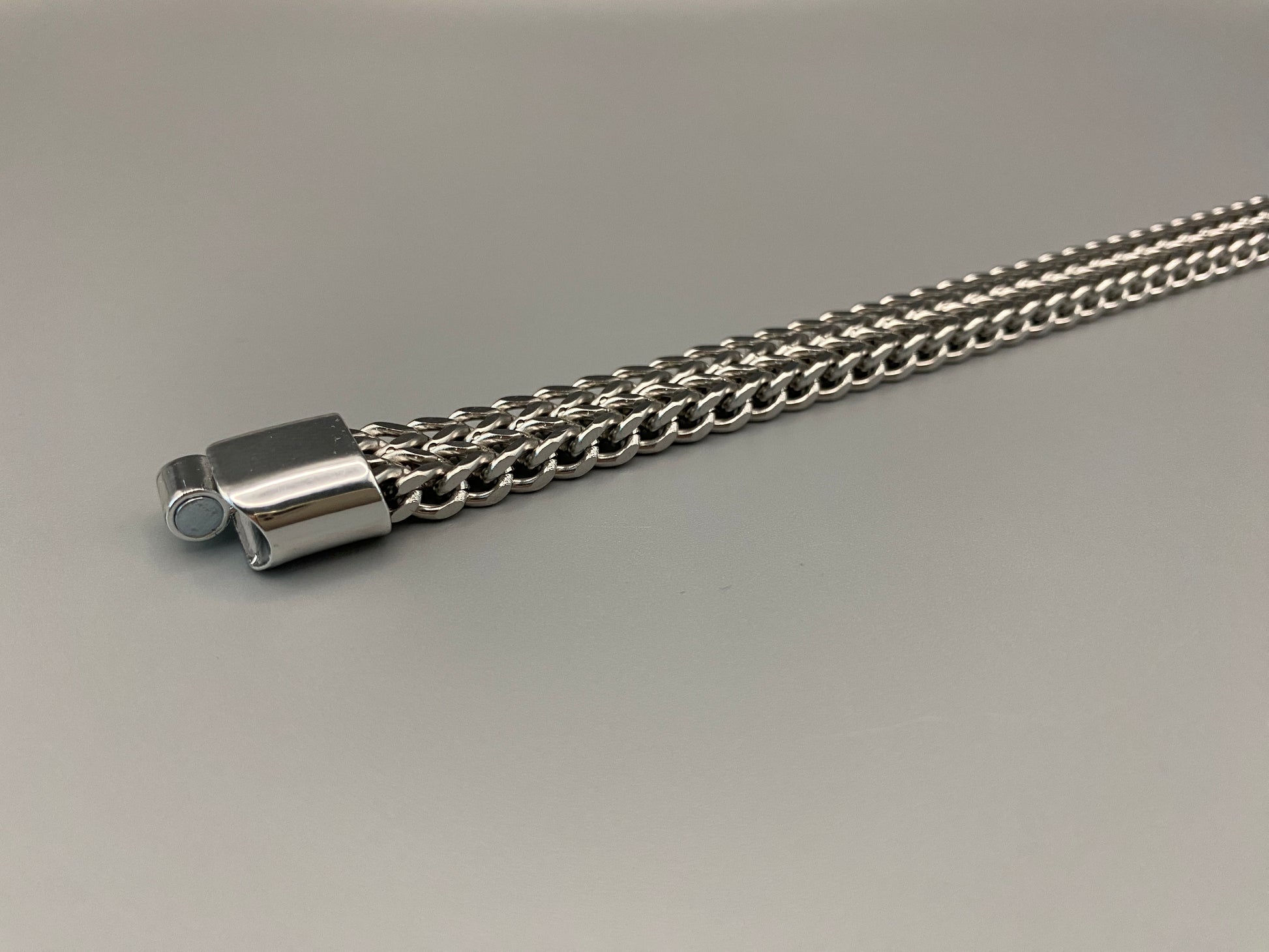 “A men’s bracelet with a chain and link design, made from shiny titanium steel. The bracelet features a geometric pattern and a smart magnet button clasp. It is available in silver, gold, and black colors, and measures 19-23 cm in length.”